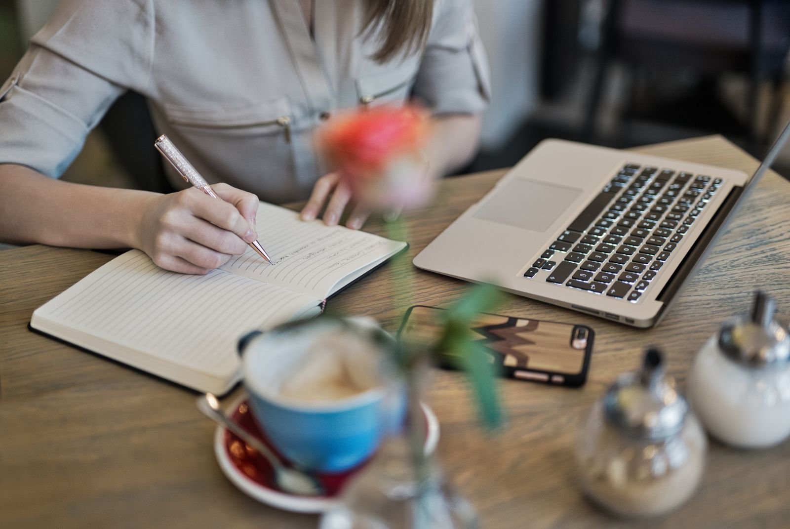 Woman writing in a notebook beside a laptop and coffee cup on a desk
